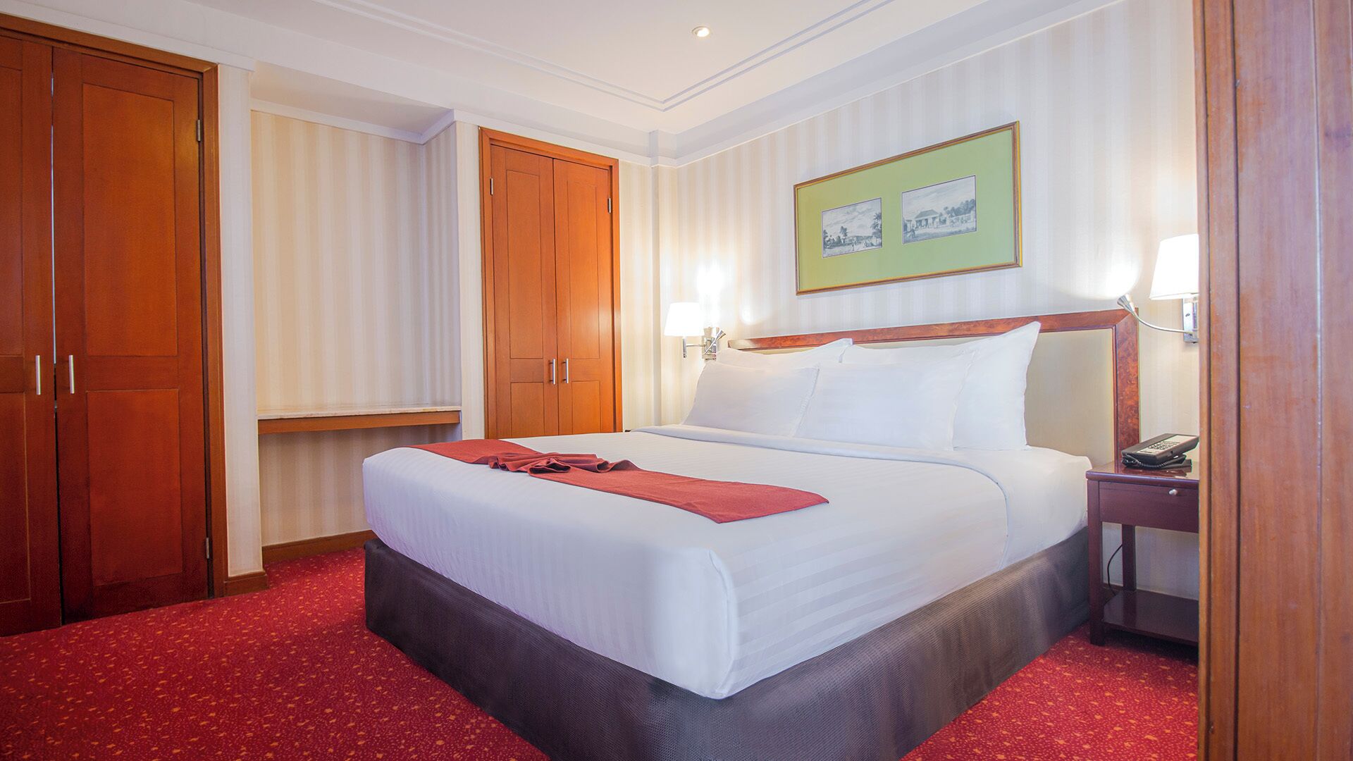 A hotel room with a large bed, a wooden wardrobe, a bedside table with a lamp, and a framed picture on the wall.