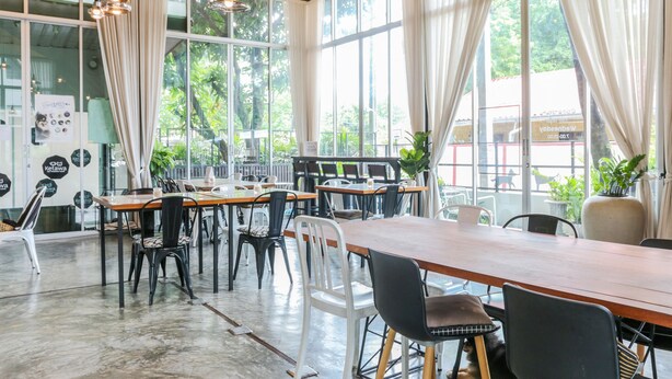 Un café amplio con ventanas grandes, mesas de madera y sillas negras y blancas.