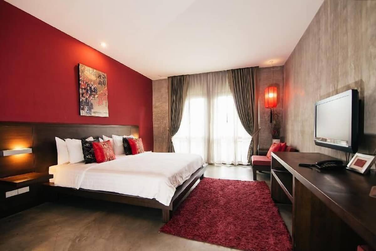 A hotel room with a bed, a desk, a TV, a red rug, and a painting on the wall.