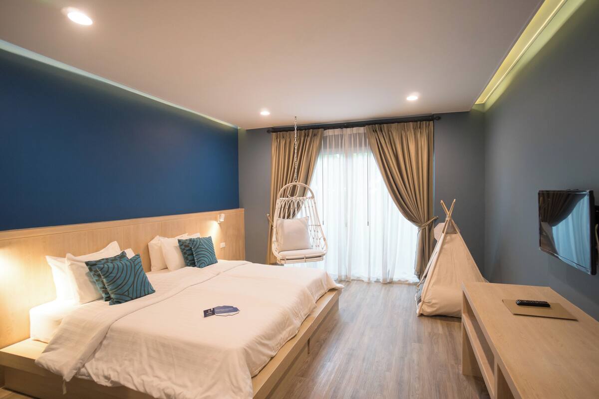 A modern hotel room with a large bed, a hanging chair, a sofa, and a TV.
