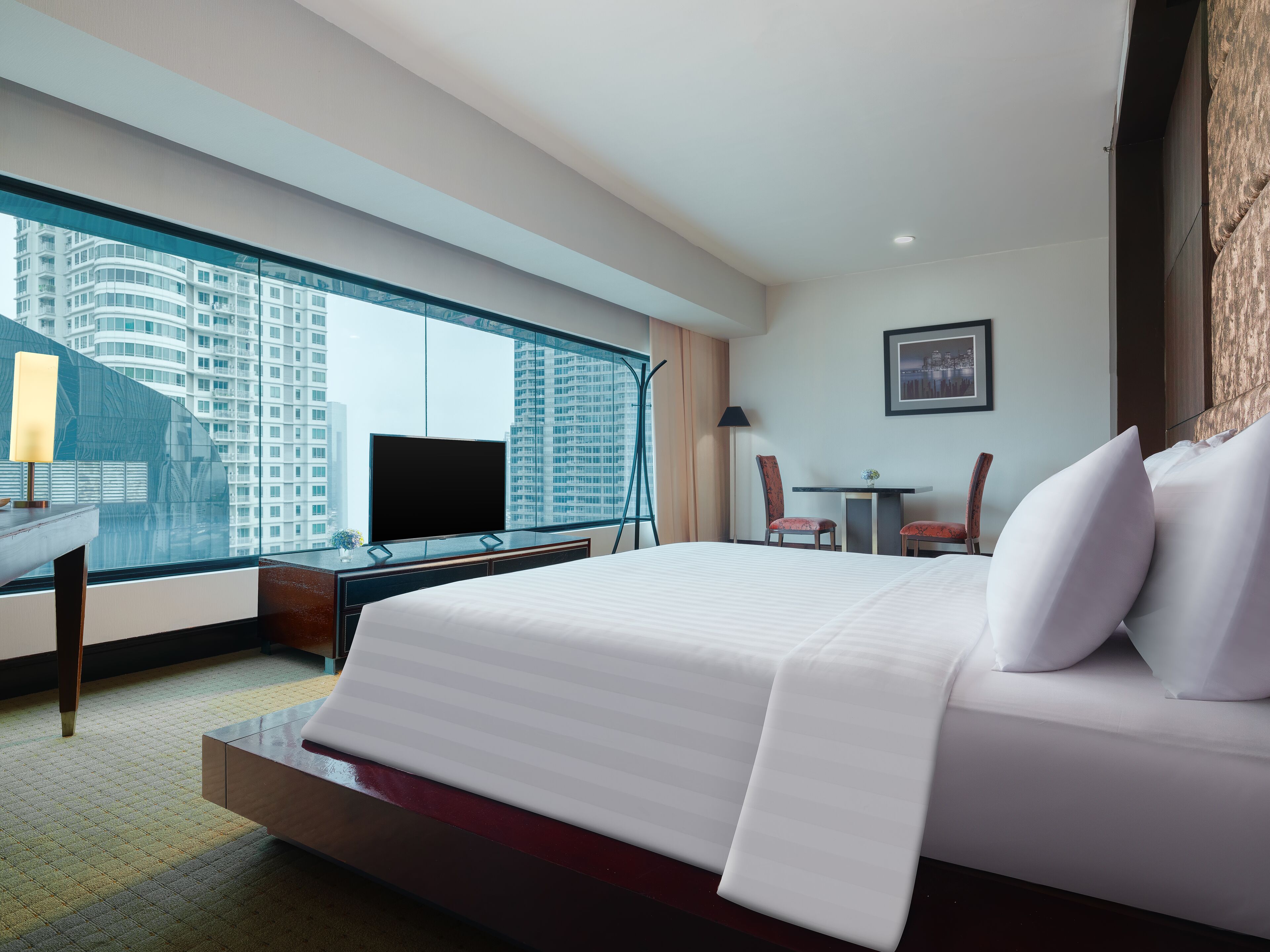 A hotel room with a large bed, a desk with a TV, a chair, a small table, and a view of a cityscape through a large window.