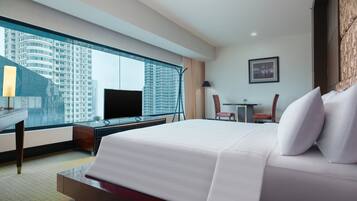 A hotel room with a large bed, a desk with a TV, a chair, a small table, and a view of a cityscape through a large window.