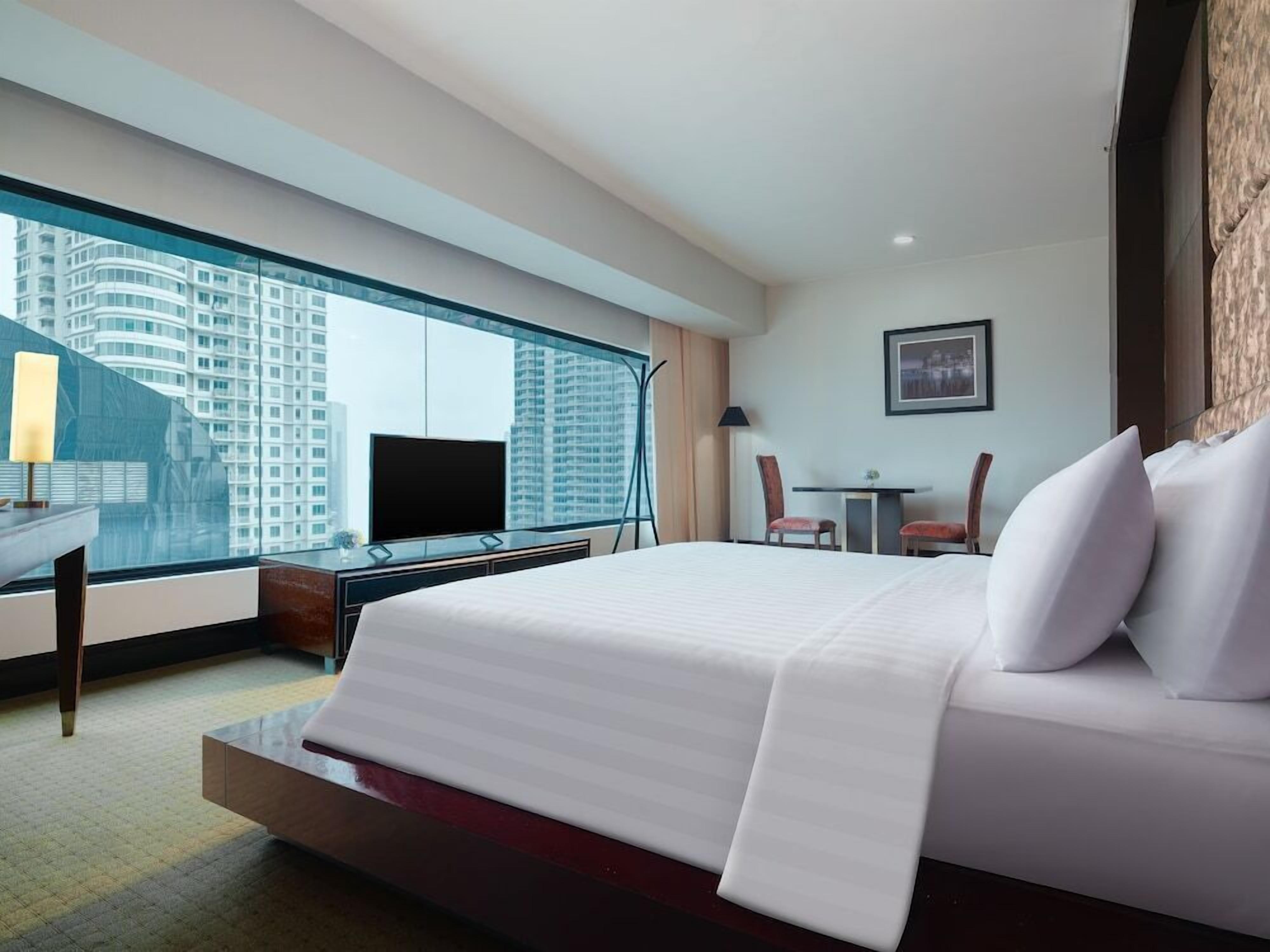 A hotel room with a large bed, a desk with a chair, a TV, and a view of high-rise buildings.