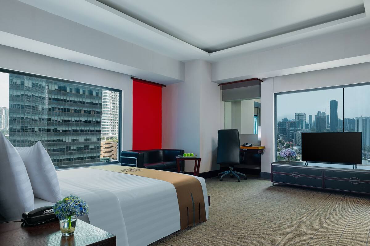 A hotel room with a bed, a desk, a TV, and a large window with a city view.