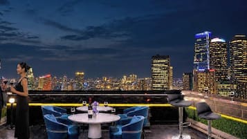 Una pareja en un bar en la azotea con vistas al horizonte urbano, mesas con sillas y botellas de vino.