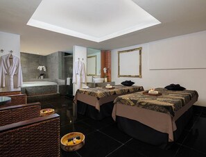 Une chambre d’hôtel moderne avec deux lits, un coin salon doté d’un fauteuil en osier, un grand miroir et une baignoire.