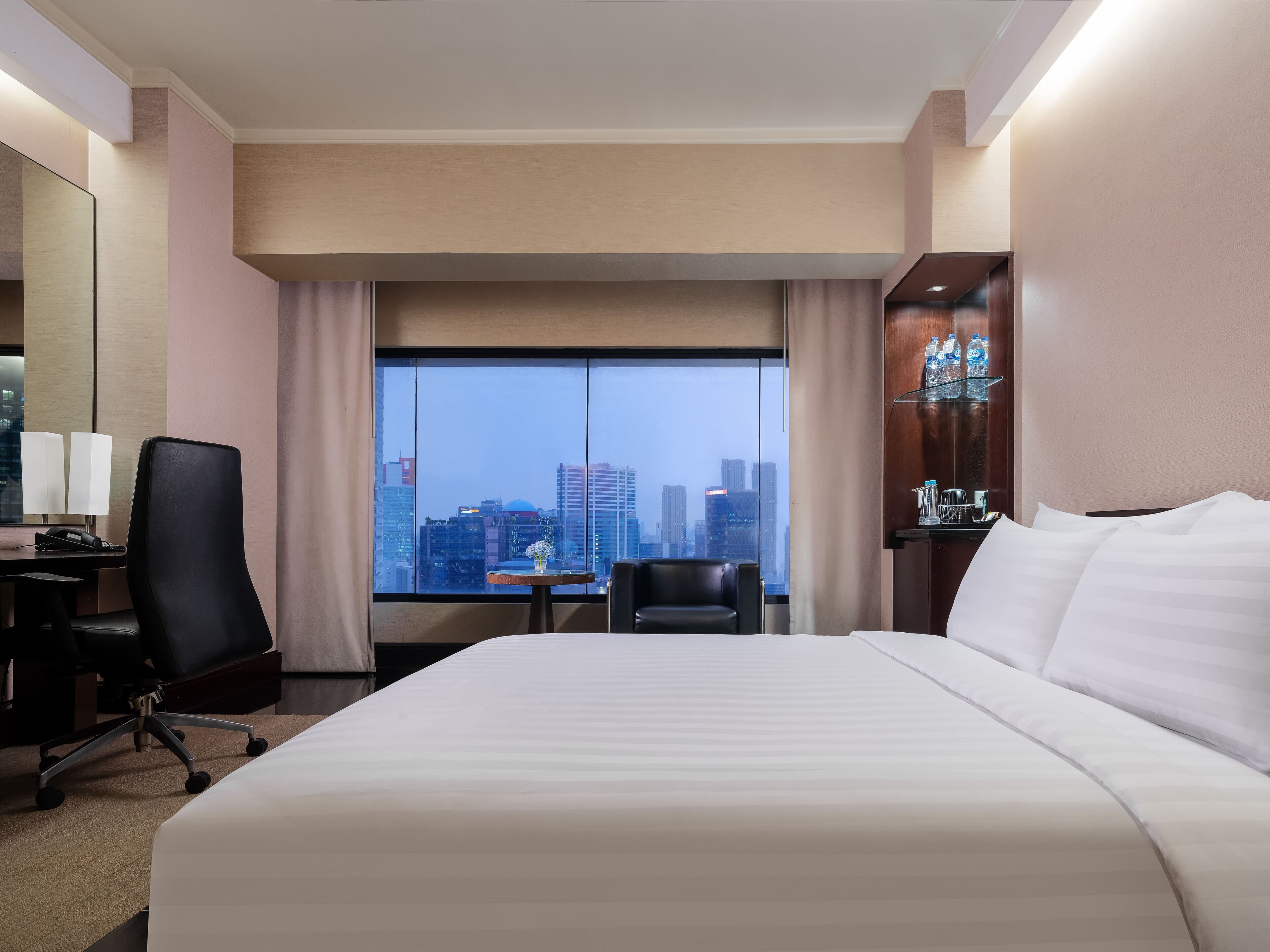 Une chambre d’hôtel avec un grand lit, un bureau avec une chaise, une petite table avec un vase, et une vue sur le paysage urbain par une grande fenêtre.