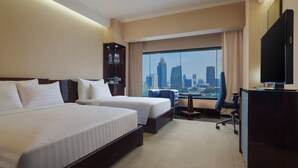 Habitación de hotel con dos camas, un escritorio, una silla, un televisor y un ventanal con vistas a la ciudad.