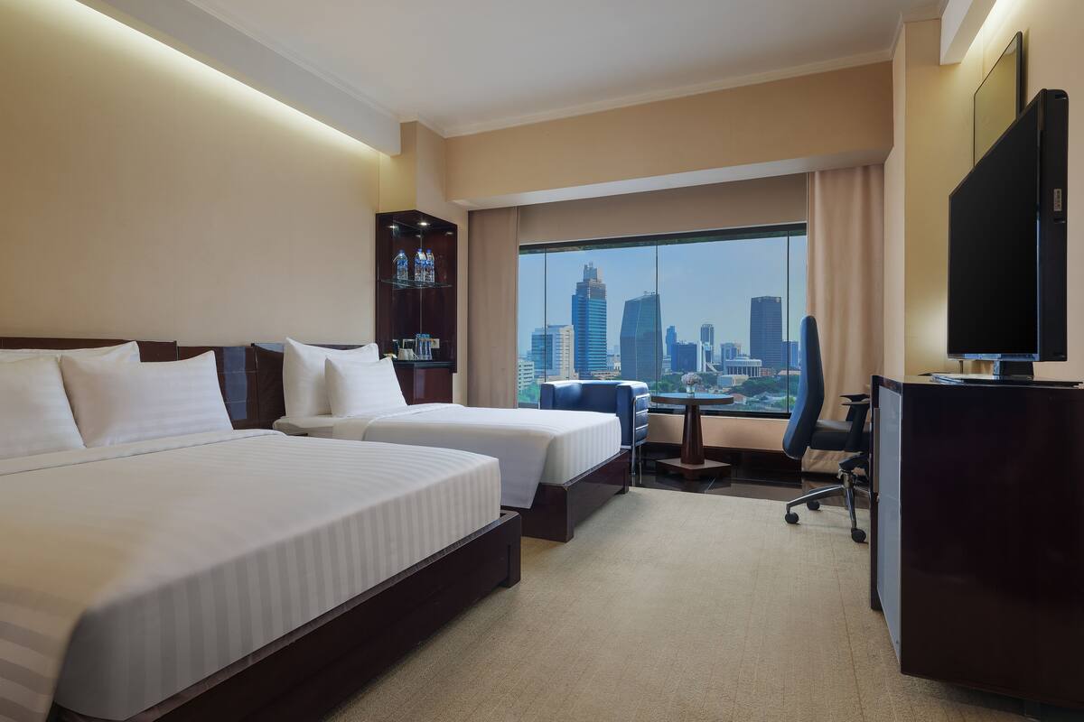 A hotel room with two beds, a desk, a chair, a TV, and a large window with a city view.