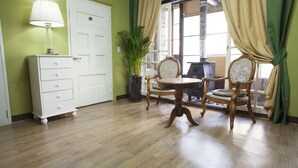 Une pièce avec un plancher en bois, une commode blanche, une lampe, deux chaises, une table, une plante en pot et une fenêtre avec des rideaux.