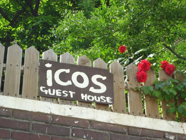 A wooden sign with the text "ICOS GUEST HOUSE" on a brick wall, with a wooden fence and red roses in the background. - iCOS Guesthouse 2 for Female - Hostel (Seoul)