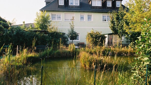 A two-story house with a sloped roof, surrounded by a pond and dense vegetation. - Hotel Gasthof Goldener Hahn (Frankfurt an der Oder)