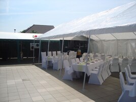 Un área de comedor al aire libre con carpas, manteles blancos y sillas, suelo de baldosas y un edificio al fondo.