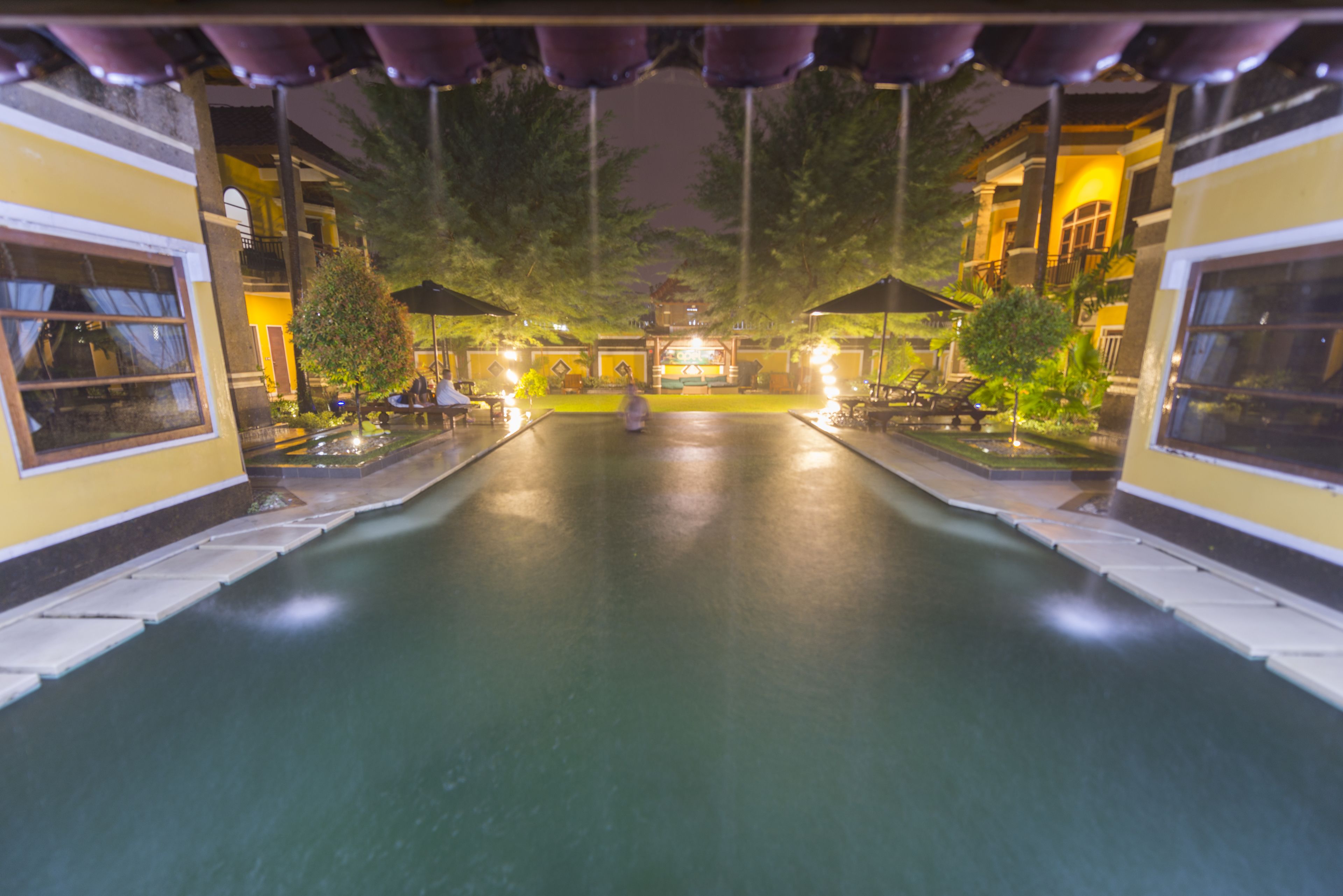 A nighttime swimming pool area with lounge chairs and umbrellas, surrounded by buildings with balconies.