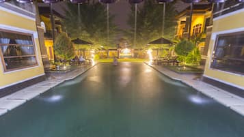 A nighttime swimming pool area with lounge chairs and umbrellas, surrounded by buildings with balconies.