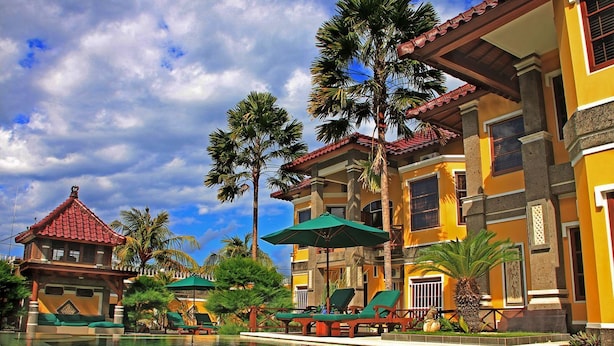 A pool area with lounge chairs and umbrellas, surrounded by palm trees and a multi-story building with a red-tiled roof. - Apel Villa Sanur (Denpasar)