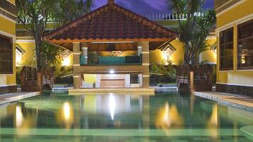 A swimming pool with a covered seating area, surrounded by palm trees and a building with a tiled roof.