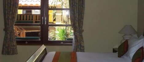 A hotel room with a bed, a window with curtains, a nightstand with a lamp, and a view of the outside.