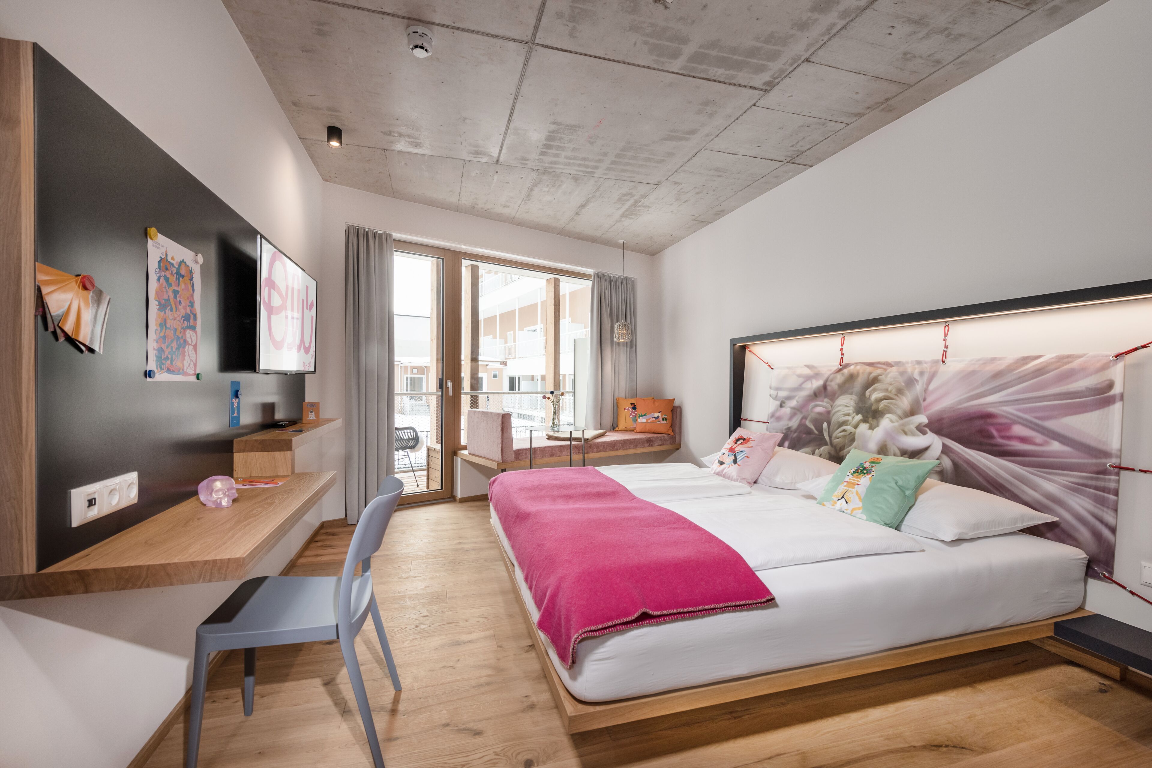 A modern bedroom with a large bed, a desk, and a chair. There is a window with curtains, a television on the wall, and artwork.