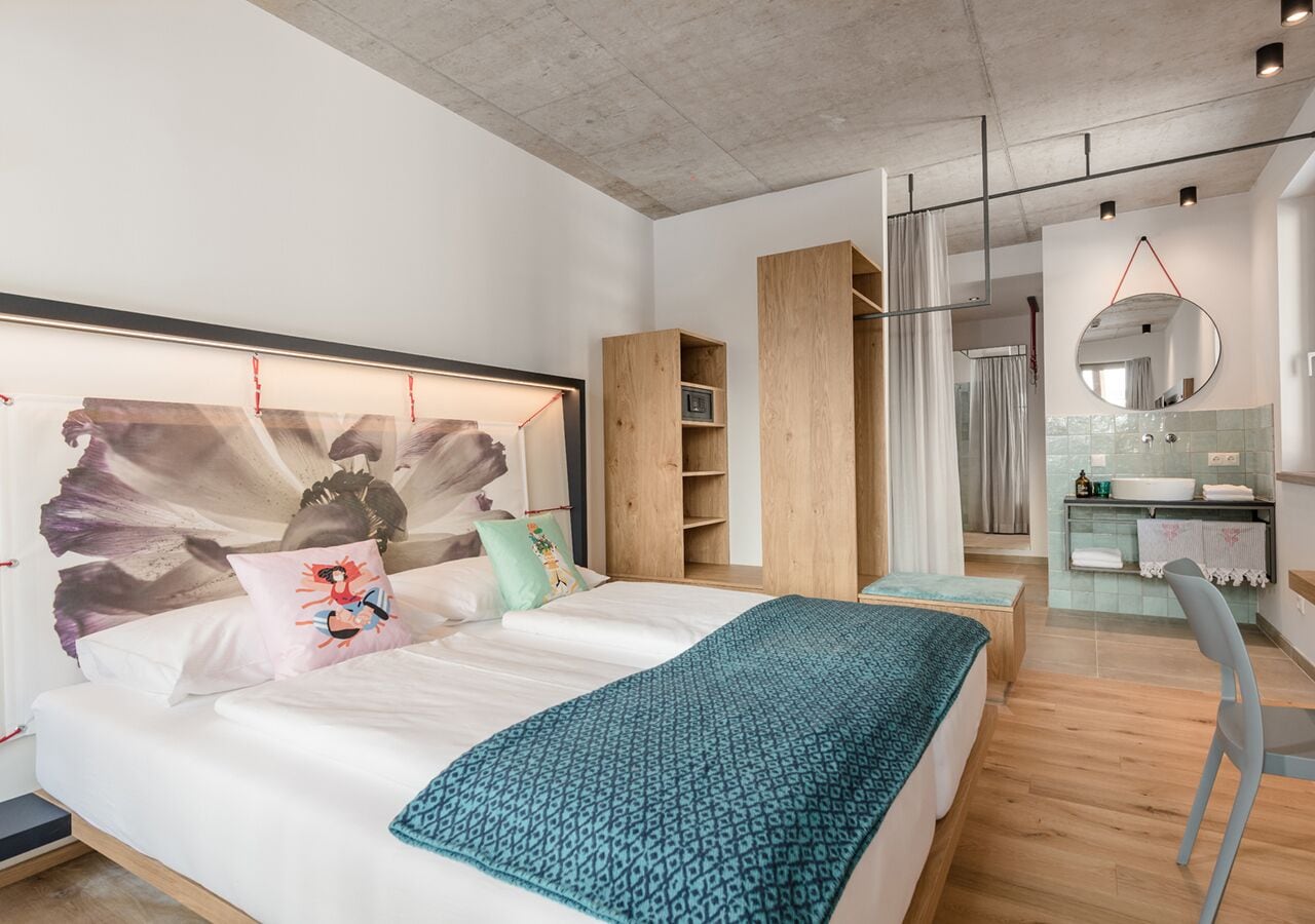 A modern bedroom with a large bed, a wooden wardrobe, a bathroom with a mirror, and a small seating area.