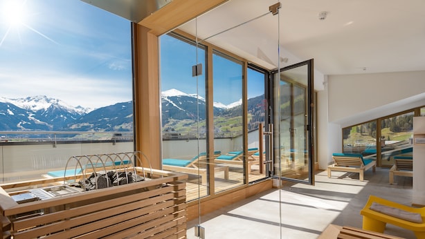 Un balcón moderno con vistas a montañas nevadas, mobiliario de madera y una barandilla de vidrio.
