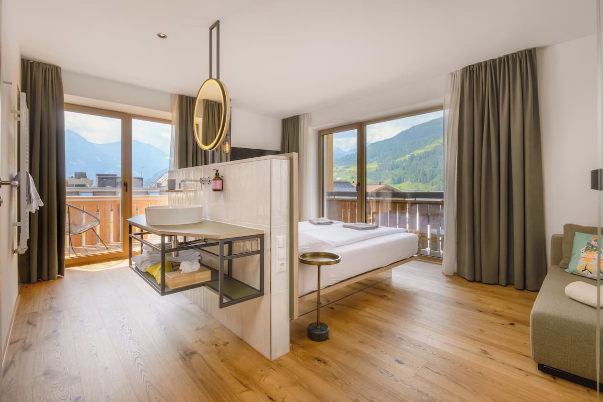 Habitación de hotel con suelo de madera, una cama, una mesita con espejo, un lavabo y un balcón con vistas a las montañas.