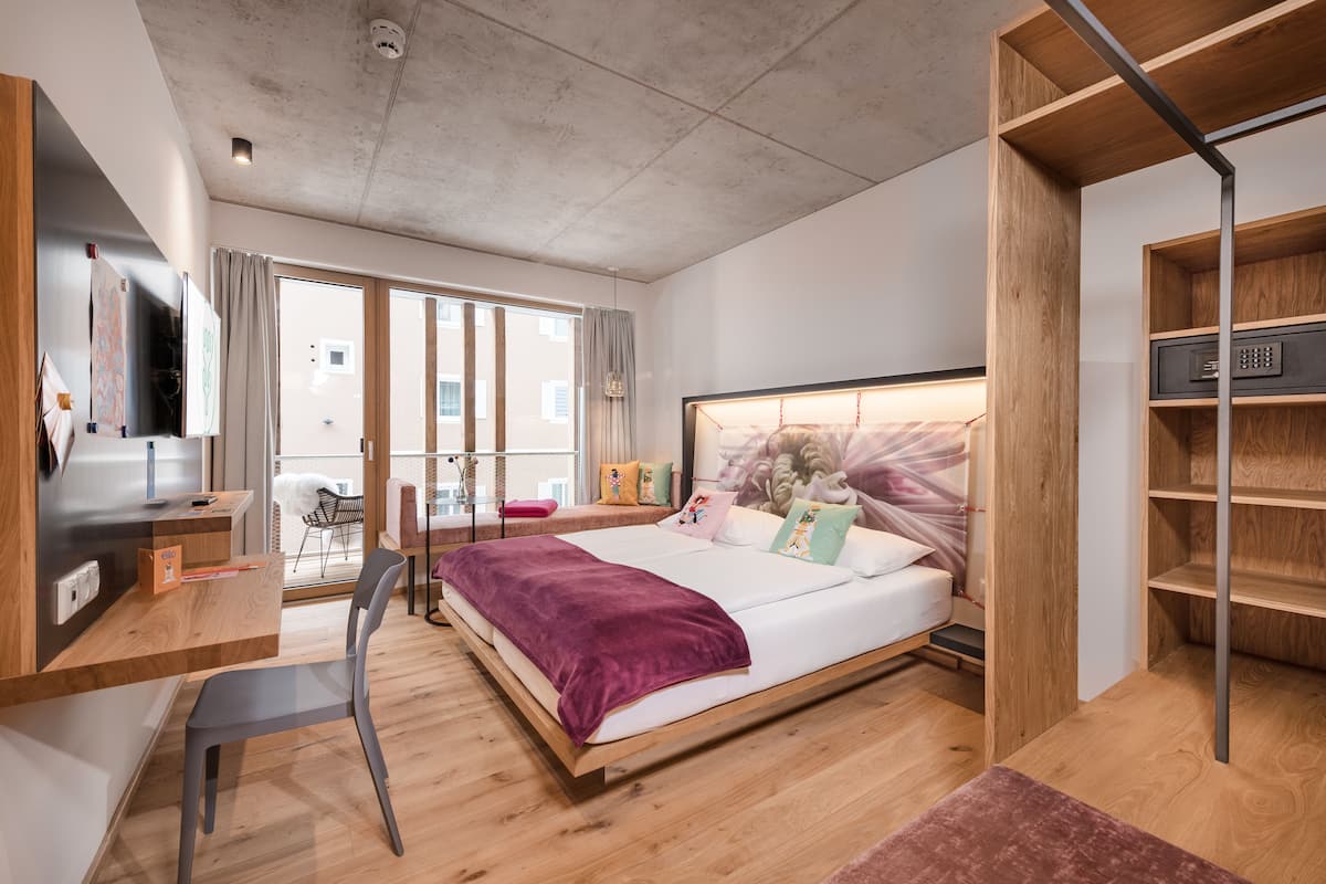 Un dormitorio moderno con una cama grande, un escritorio y una silla. Se ve una ventana con cortinas, vistas al exterior y un estante de madera.