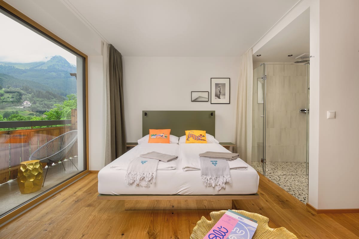 Cama doble con sábanas blancas, dos almohadas y una mesita de noche con un libro. Un amplio ventanal ofrece vistas a un paisaje montañoso.