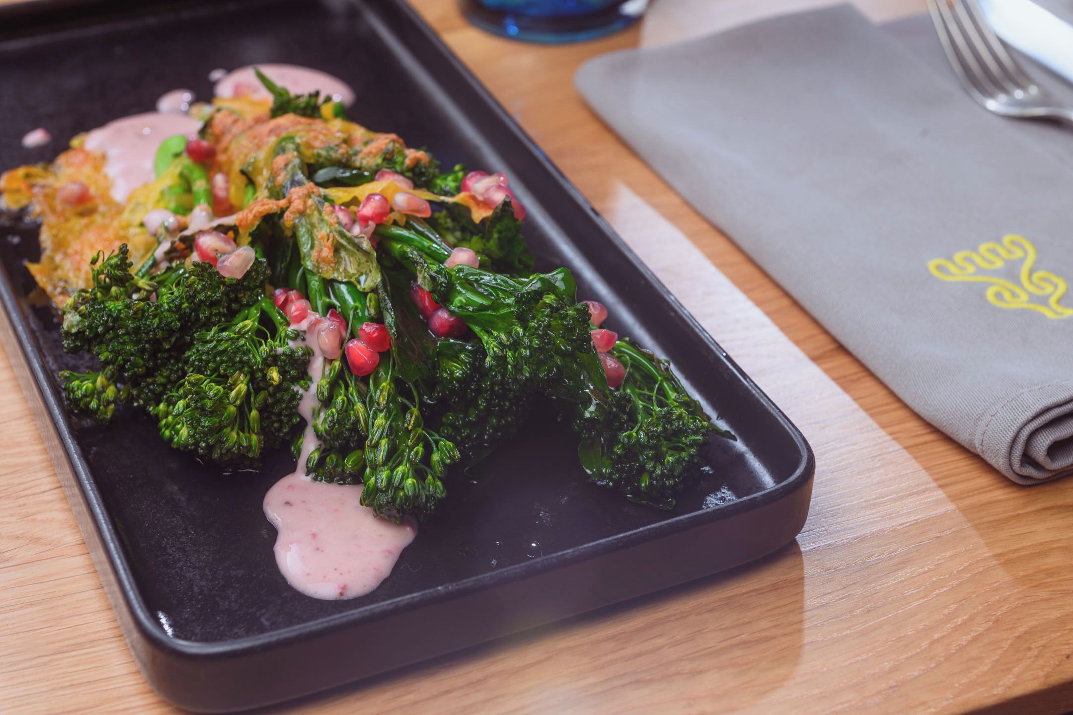 A black tray with food, including broccoli and pomegranate seeds, on a wooden table.