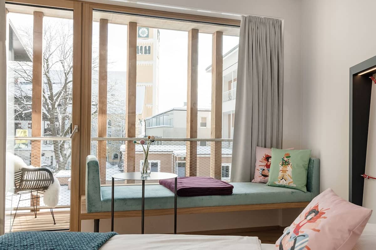 Una habitación con un ventanal grande, una cama, una mesita pequeña y un banco con cojines.