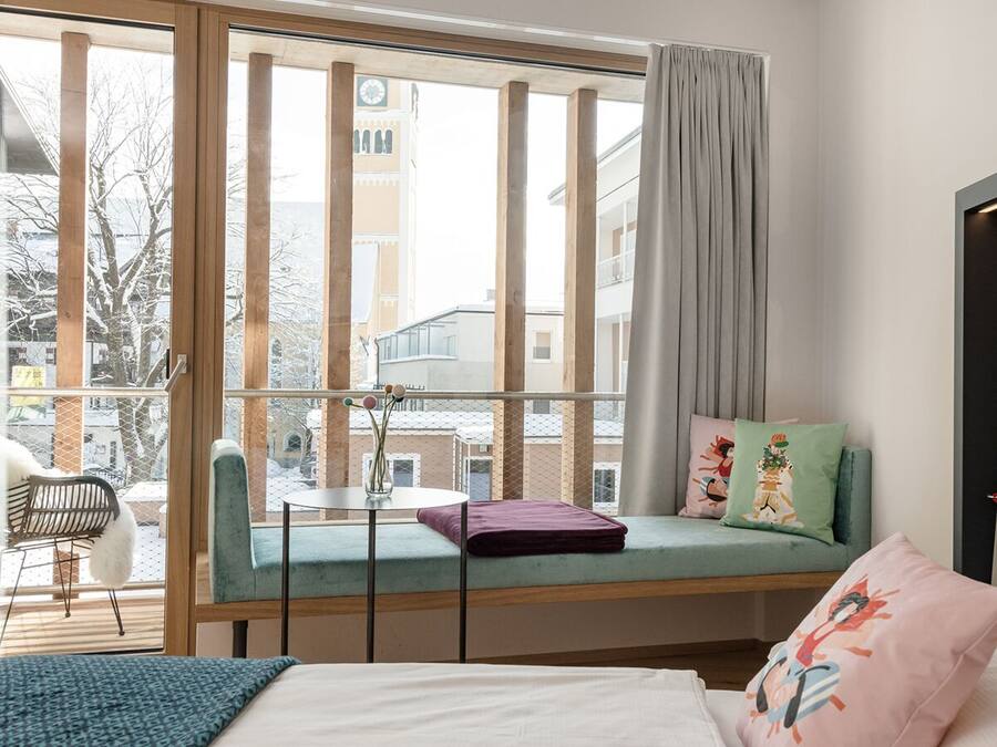 Una habitación con un ventanal grande, una cama, una mesita pequeña y un banco con cojines.