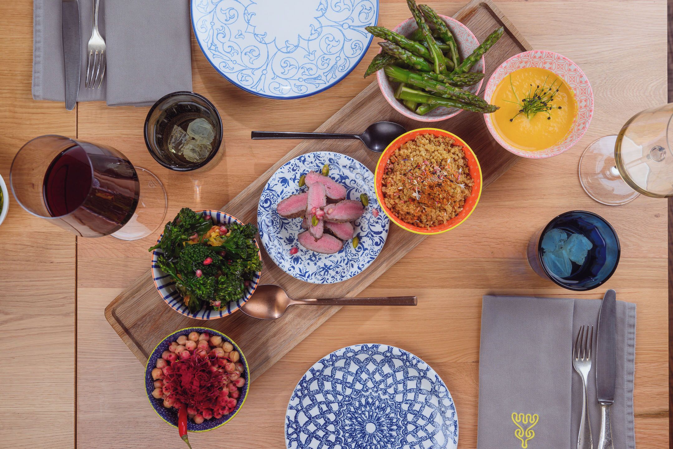 Una mesa de madera con varios platos, entre ellos un tazón de espárragos, un bol de sopa y un plato de carne. También hay copas de vino y una servilleta con cubiertos.