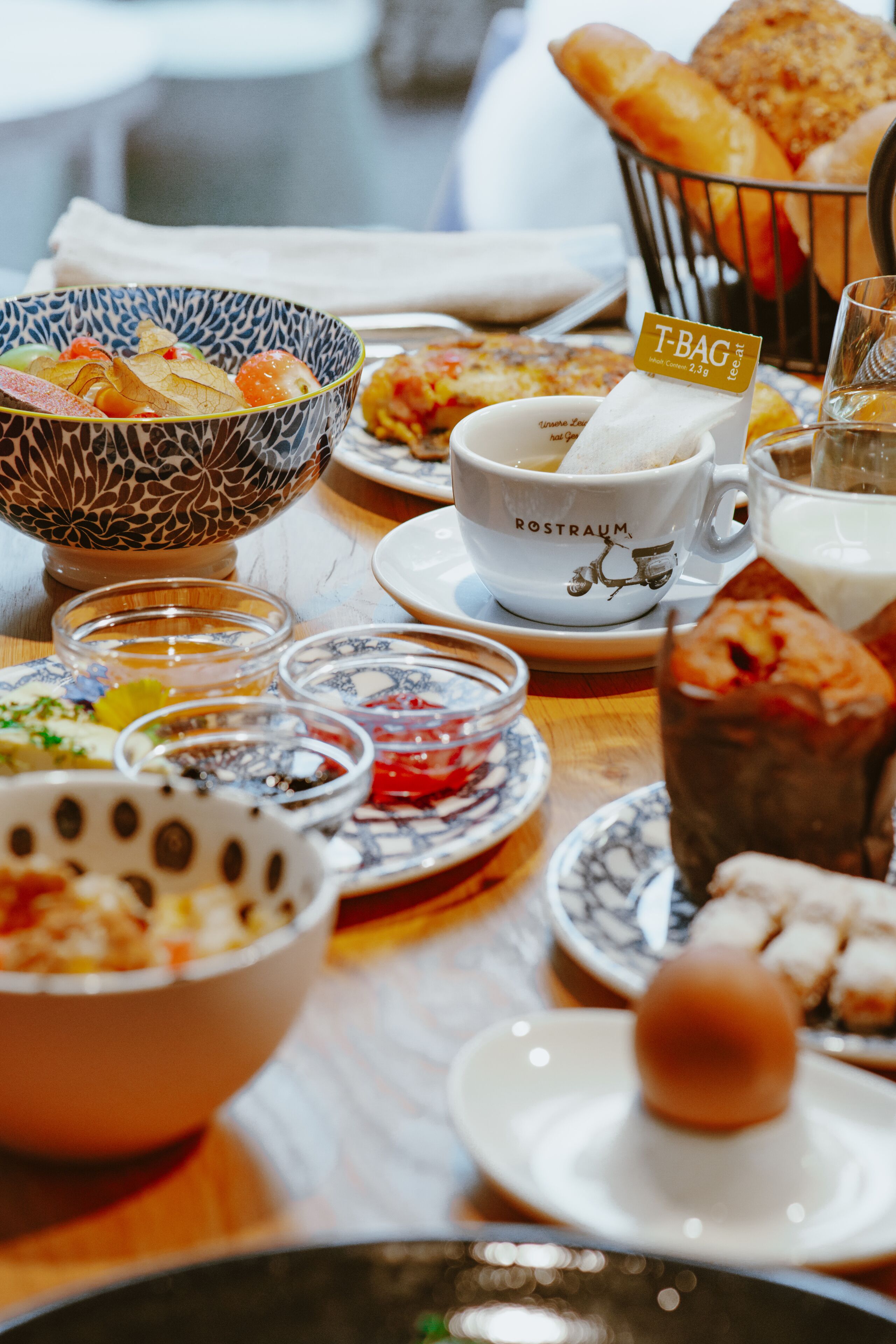 Una mesa servida con diversos platos, incluyendo un cuenco de fruta, una taza con la etiqueta "ROSTRAUM" y una cesta de pan.