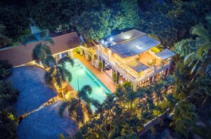An aerial view of a resort with a swimming pool, a covered outdoor area, and lush tropical vegetation.