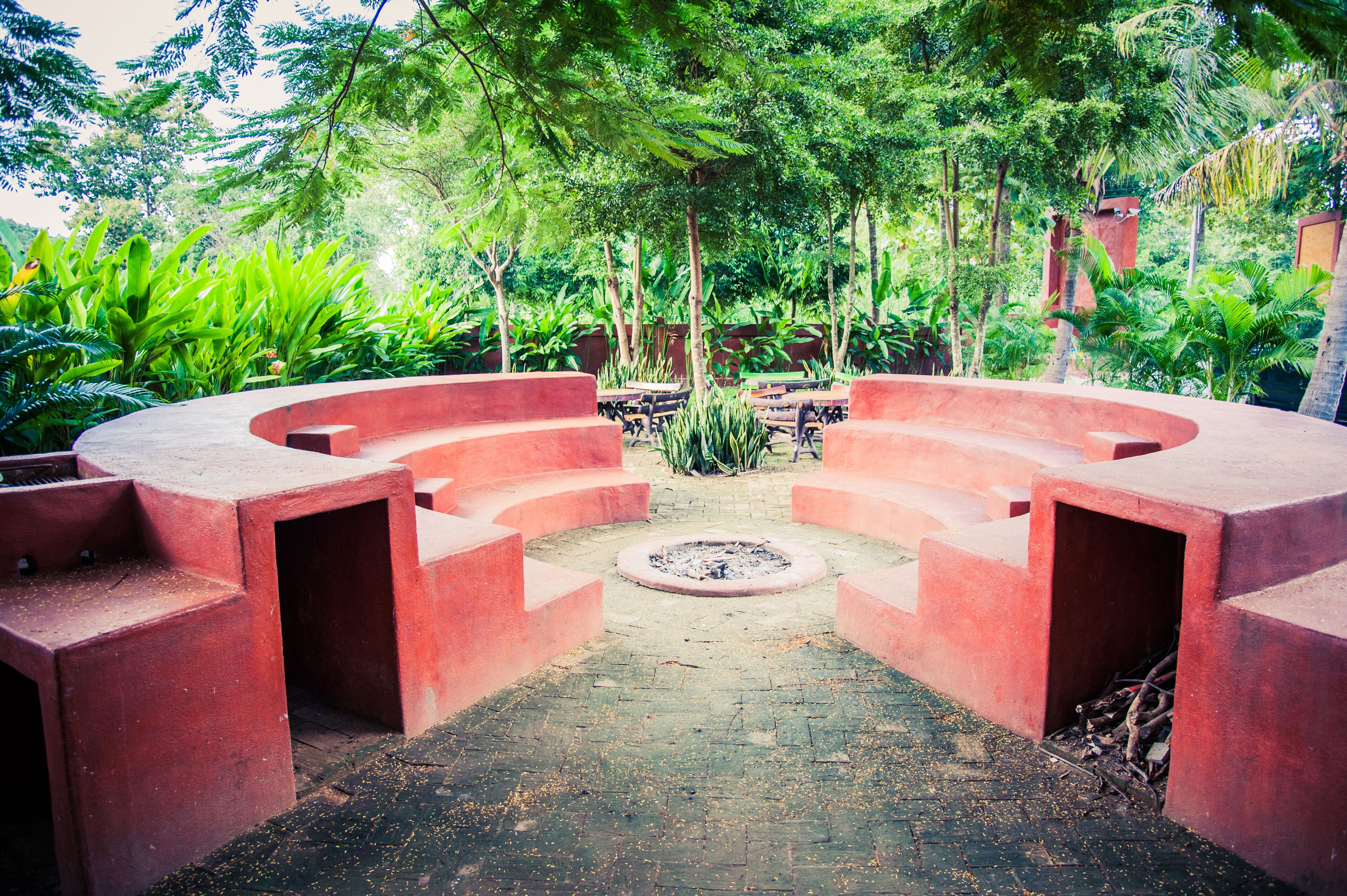 A circular outdoor seating area with red benches and a central fire pit, surrounded by lush greenery.