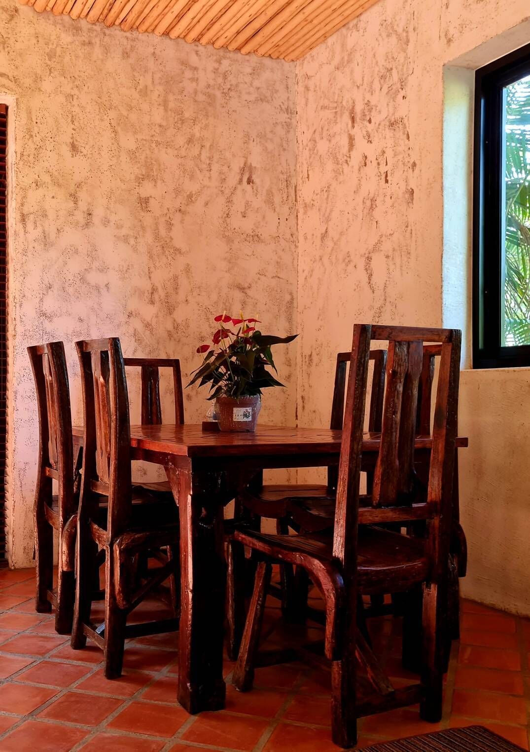 A rustic dining area with a wooden table and chairs, a potted plant, and a window with a view of greenery.