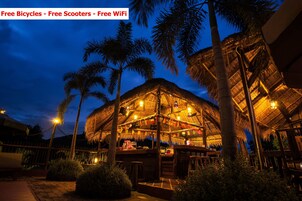Un área de comedor al aire libre con techo de paja, palmeras y luces colgantes, rodeada por una cerca de madera.