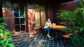Een vrouw zit aan een tafel in een beschaduwd buitengebied met een betegelde vloer, omringd door groen.
