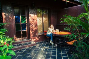 A woman sitting at a table in a shaded outdoor area with a tiled floor, surrounded by greenery.