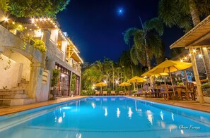 A nighttime pool area with a clear pool, outdoor seating, and a building with stairs and plants.