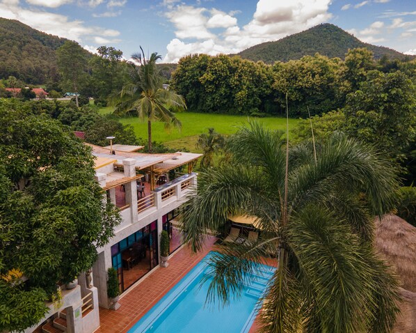 A resort with a pool, a dining area, and a view of the surrounding greenery and hills.