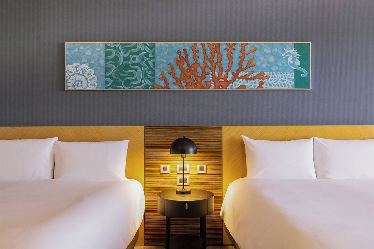 A hotel room with two beds, a wooden headboard, a wall-mounted artwork depicting coral and sea life, and a bedside table with a lamp.