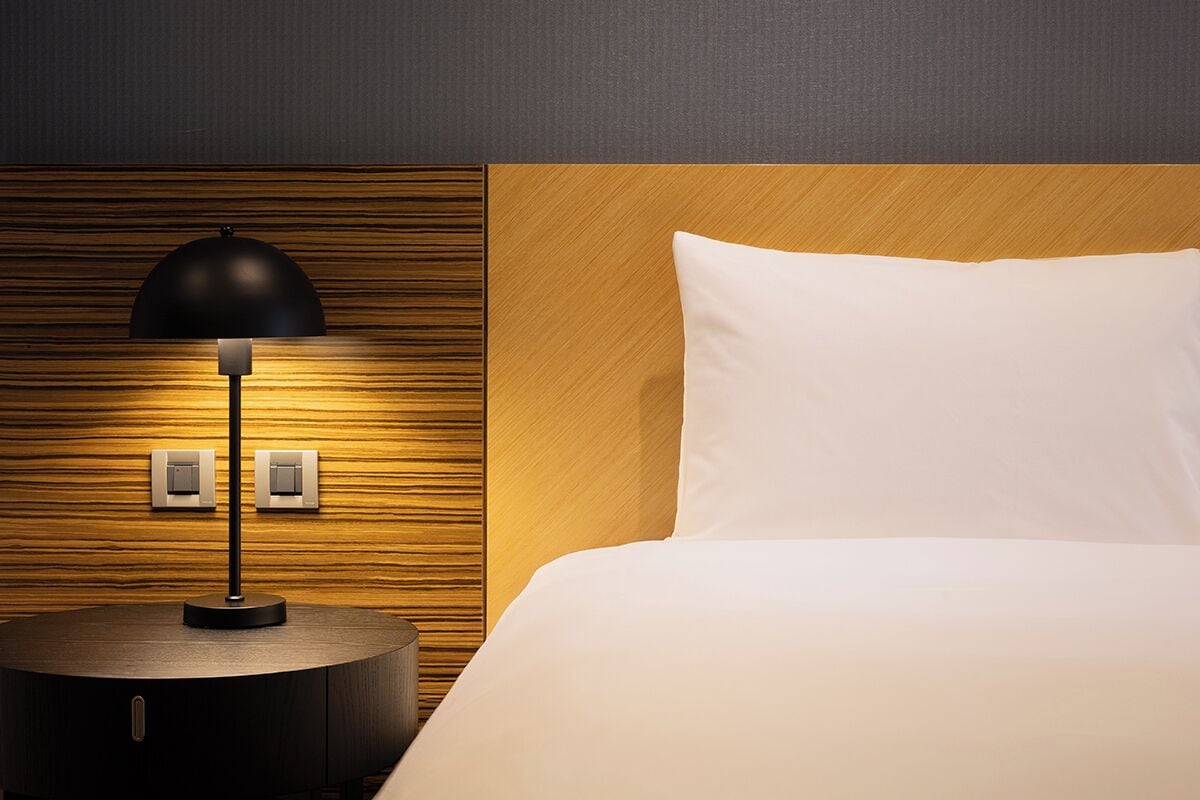 Una habitación de hotel con una mesita de noche de madera redonda, una lámpara negra, una cama blanca con cabecero de madera y dos interruptores de pared.