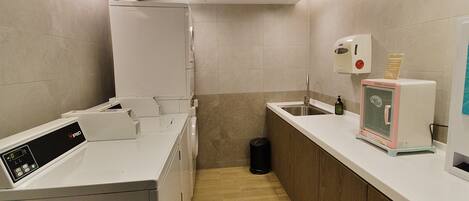 A compact hotel bathroom with a washing machine, a sink, and a hand sanitizer dispenser.