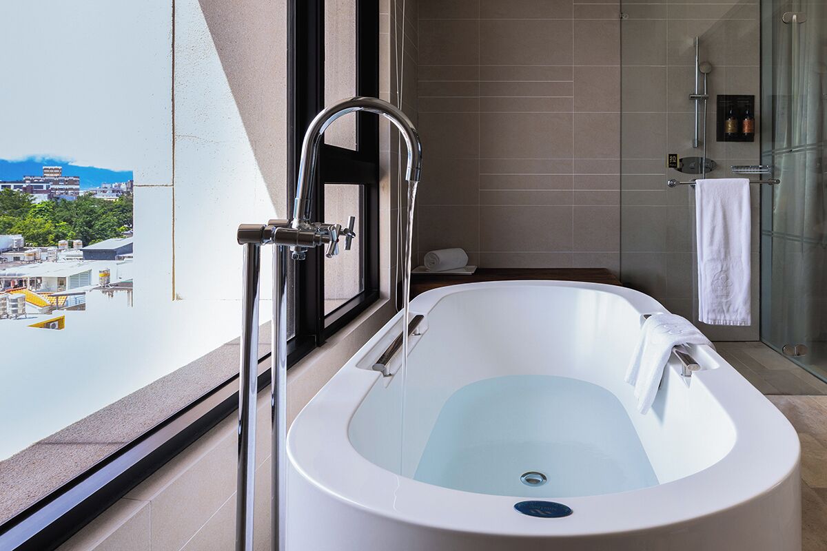 A modern bathroom with a large bathtub, a glass shower enclosure, and a view of the cityscape through a large window.