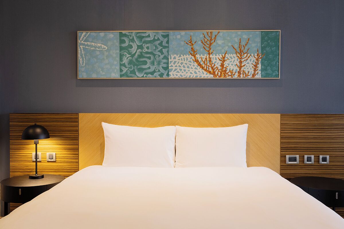 A hotel room with a bed, a nightstand with a lamp, a wall-mounted painting, and a control panel on the wall.