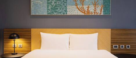A hotel room with a bed, a nightstand with a lamp, a wall-mounted painting, and a control panel on the wall.