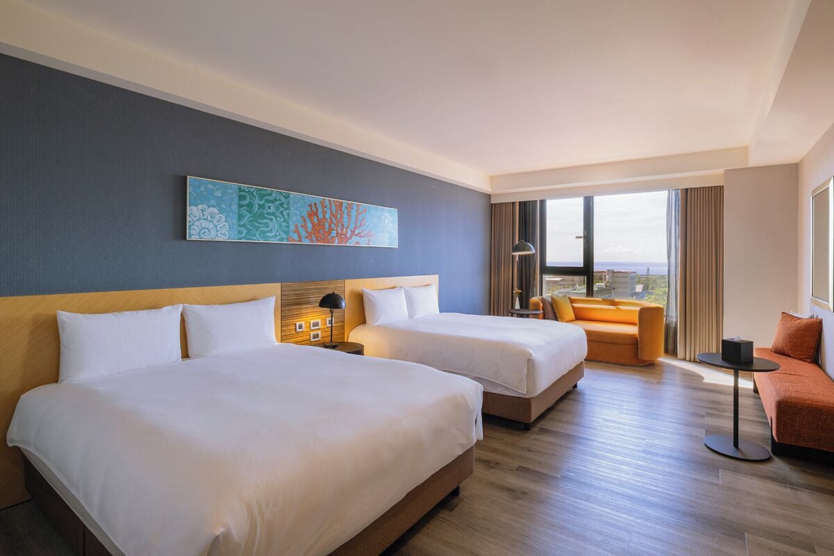 A hotel room with two beds, a sofa, a small table, and a large window offering a view.