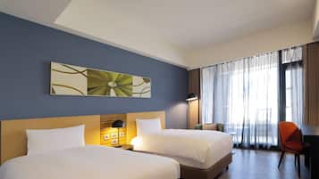 Quarto de hotel com duas camas, uma mesa, uma cadeira e uma janela grande com cortinas.