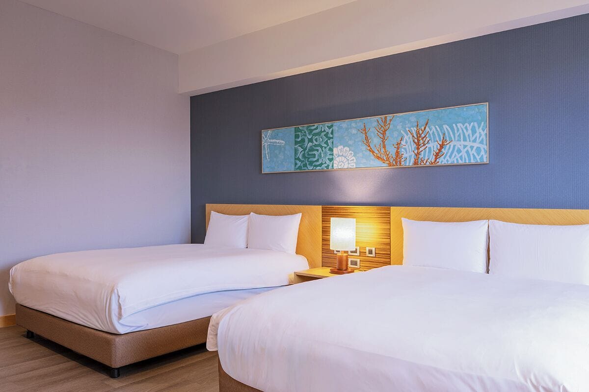 A hotel room with two beds, a nightstand with a lamp, and a wall-mounted artwork.